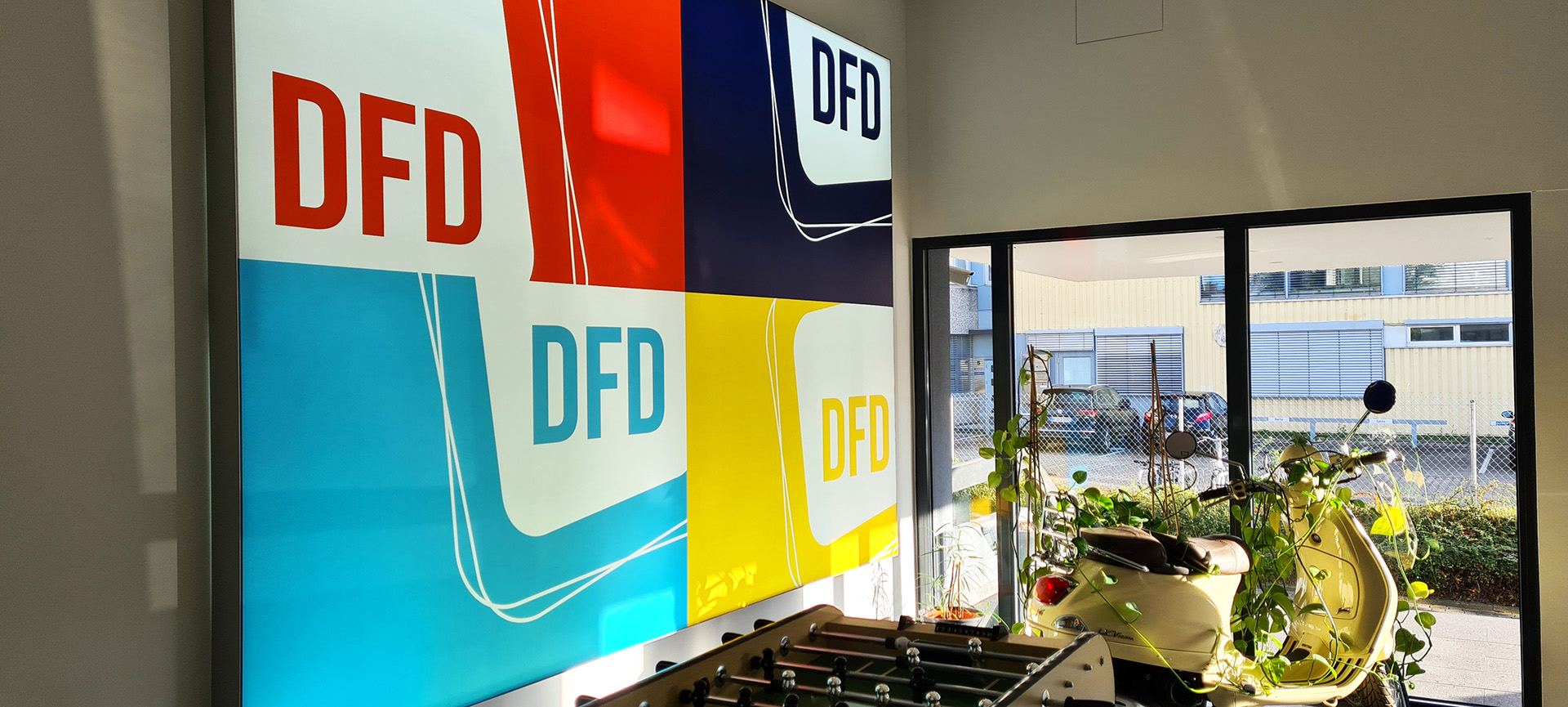 DFD-Logo an einer Bürowand mit Blick auf eine Vespa und Außenbereich.  Das Bild zeigt die moderne Arbeitsumgebung von DFD.